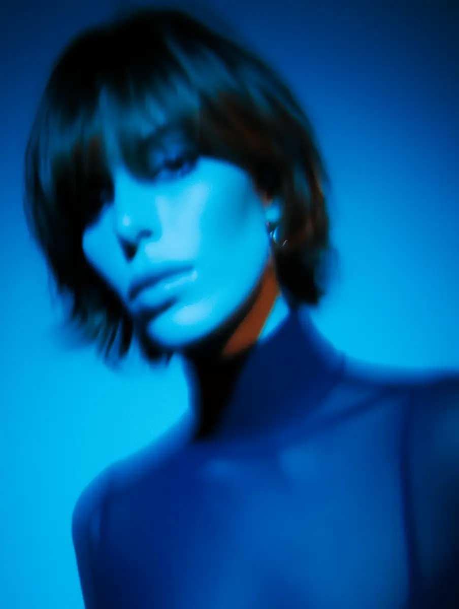 Blurred portrait of a person with short hair under blue lighting, wearing a high-neck top.